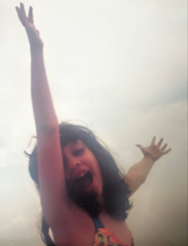 Photo of a young girl, around 8 years old, with long brown hair in a bathing suit with her hands in the air. 